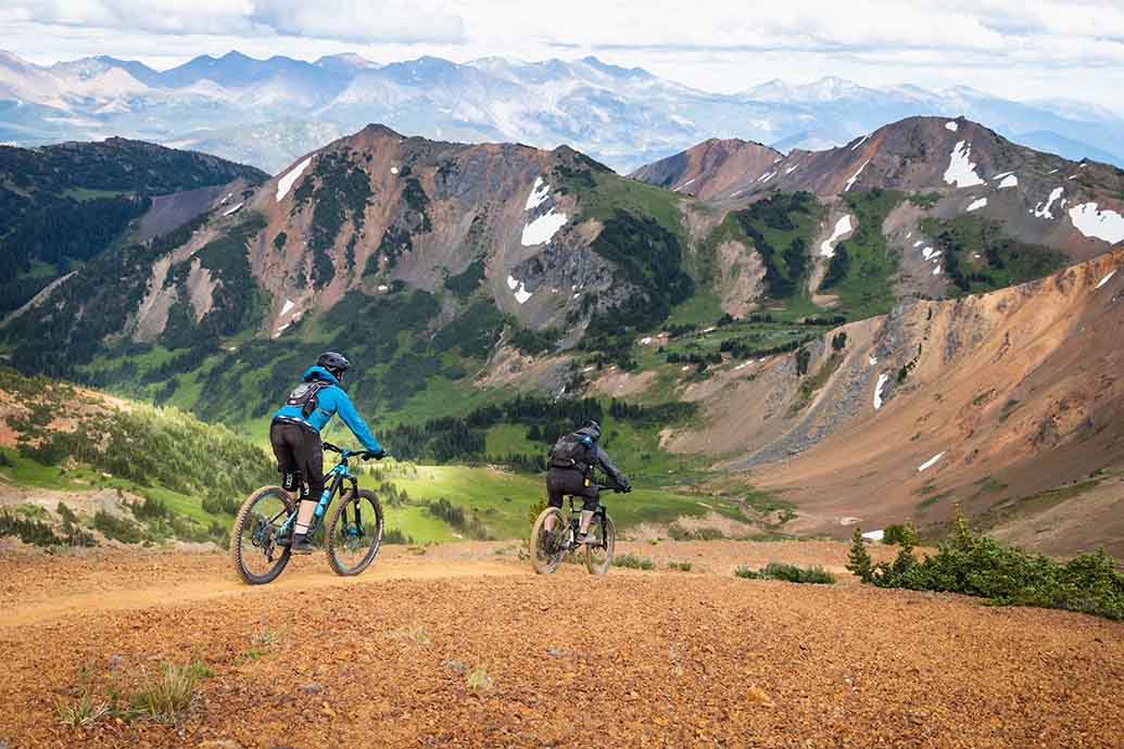 Two people biking across a mountain with a vast mountain range in the distance.